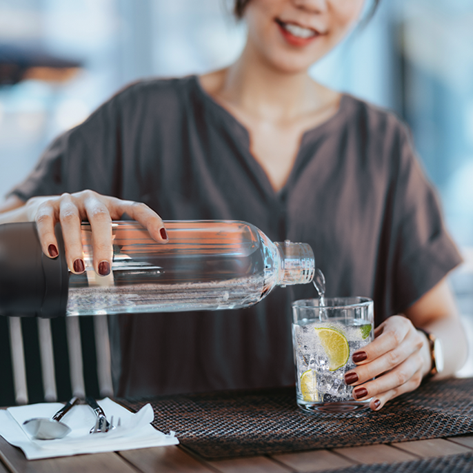 Smiling woman pouring sparkling water from a clear plastic bottle into a glass with lemon slices and ice, seated at a table with sunglasses and a napkin.