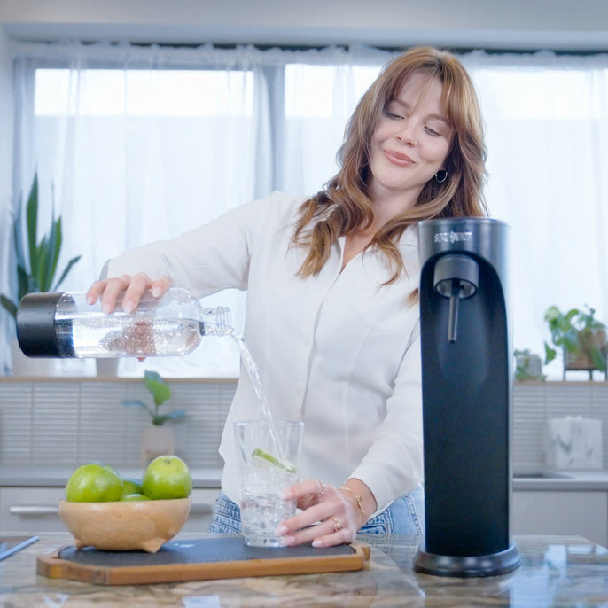 Person pouring sparkling water from a Soda Sense bottle into a glass with lime slices in a bright, modern kitchen. A black Soda Sense soda maker and a wooden bowl of fresh limes are on the counter.