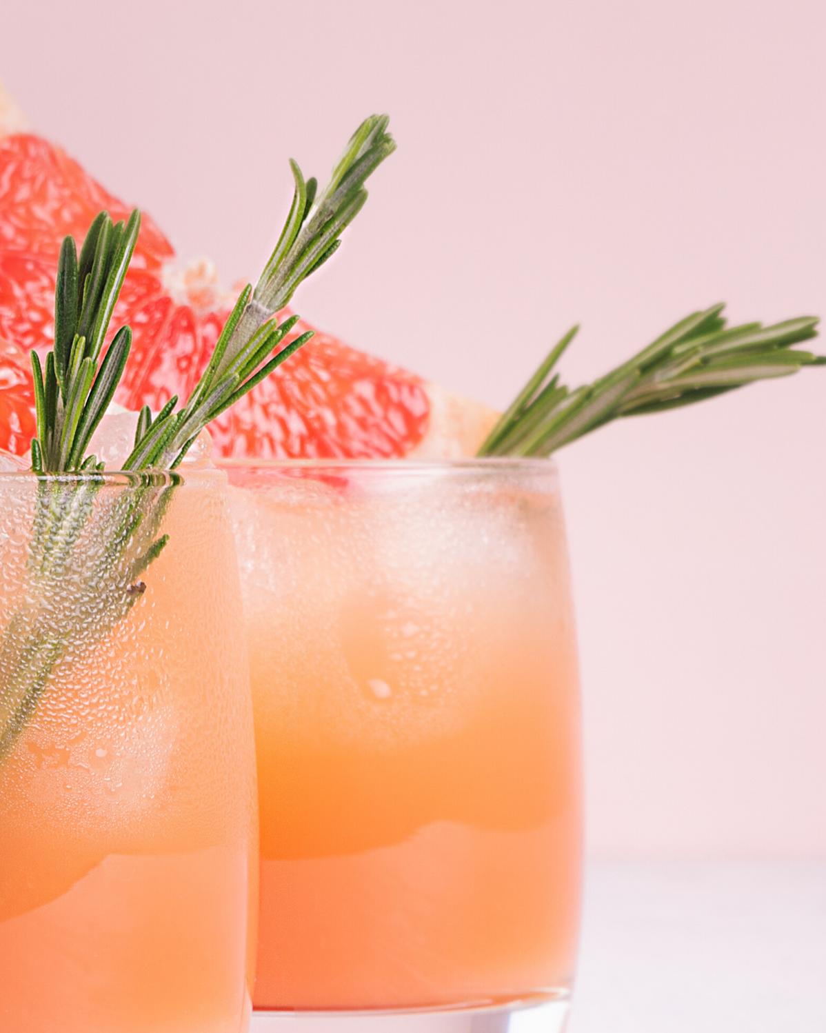 Two glasses of a pink grapefruit cocktail with ice, garnished with fresh rosemary sprigs and grapefruit slices, set on a light pink background.